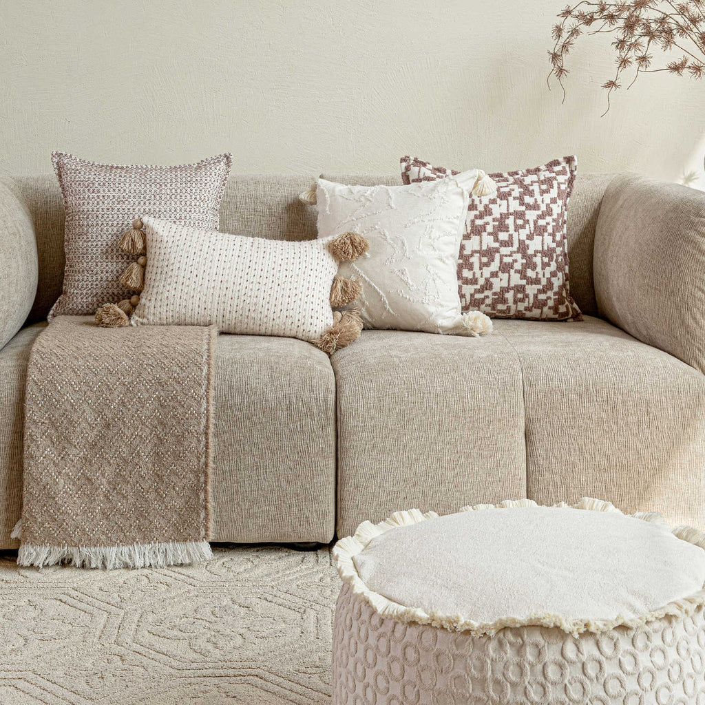 Lifestyle view of layered cotton blend cushions and a tufted ottoman in a warm, minimalist living room.